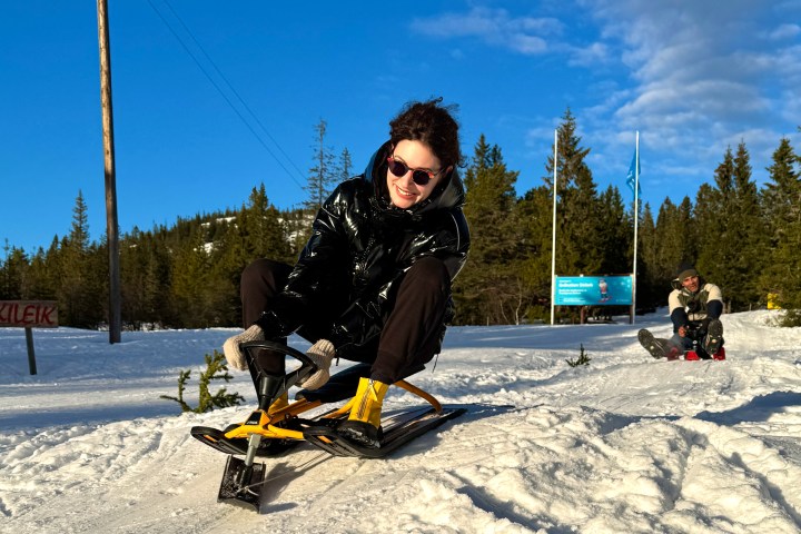 Person sledding downhill on a snowy slope in sunny weather, wearing sunglasses.