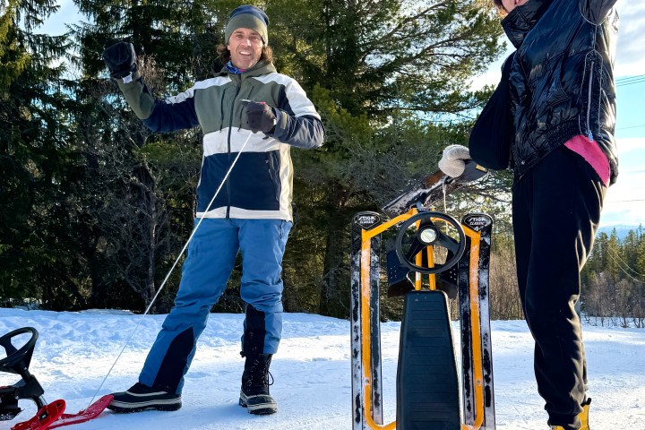 Two people in winter gear enjoying snow with a sled, tall trees in the background, sunny day.