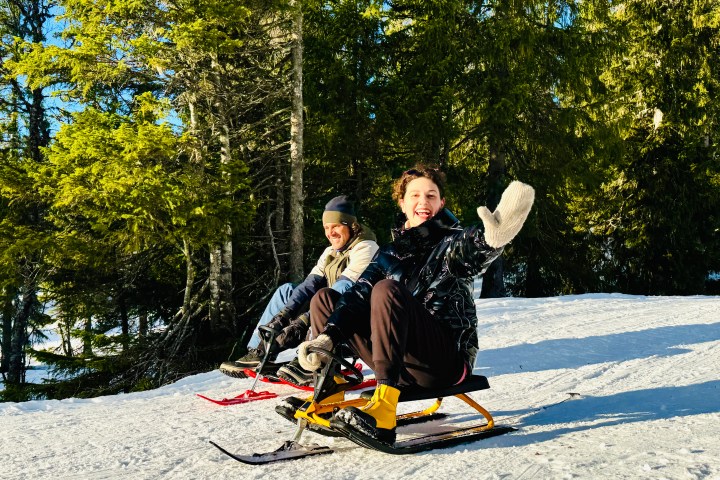 Two people sledding in a snowy forest, waving and smiling under a clear blue sky.