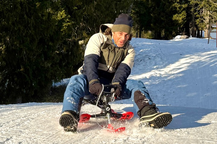 Person sledding down snowy hill in winter attire, surrounded by trees.