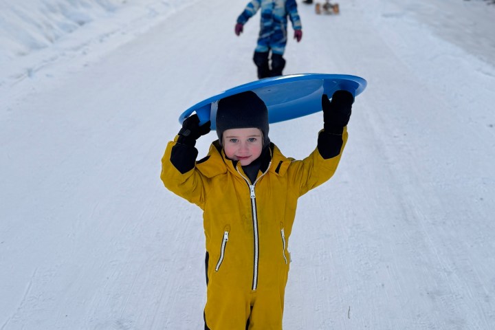Child in yellow snowsuit holds blue sled on snowy forest path, another child and trees in background.