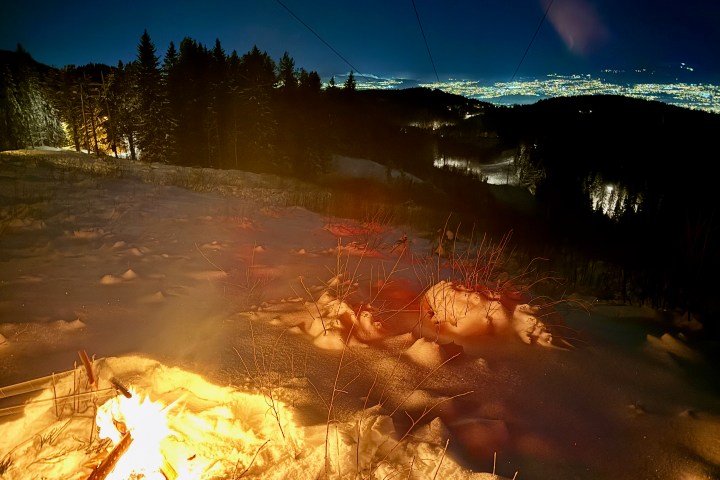 Nighttime snowy landscape with a campfire, trees, and distant city lights.