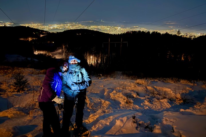 Two people stand on a snowy hill at night with city lights in the background.