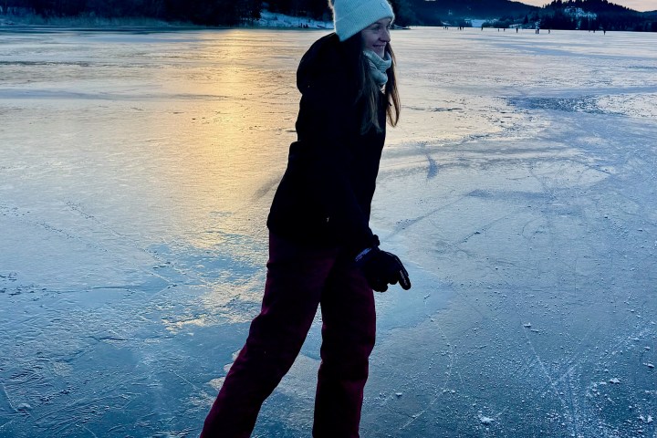 Person ice skating on a frozen lake at sunset, wearing a beanie and winter clothing.