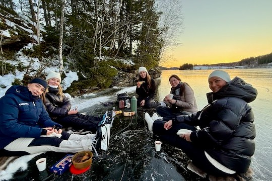 Five people sitting on ice by a lake at sunset, enjoying hot drinks.