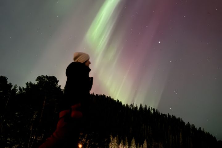 Person skating on ice under vibrant green and pink aurora borealis with forest and buildings nearby.