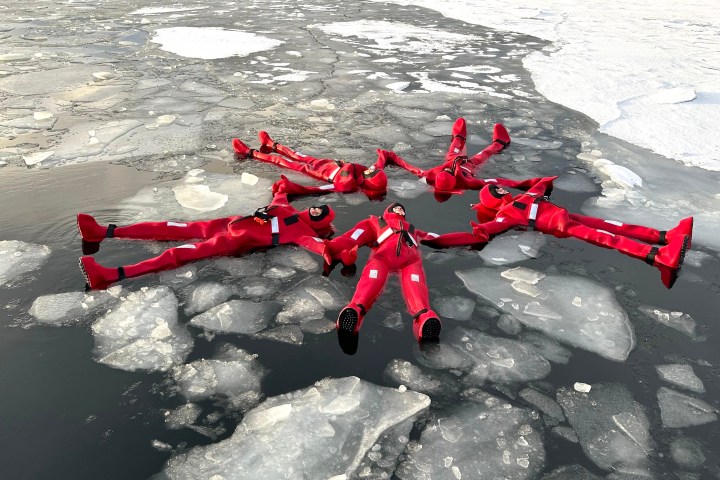 Five people in red suits floating on icy water, surrounded by ice floes.