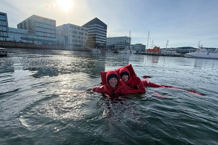 Two people in red suits floating in a harbor with buildings and boats in the background.