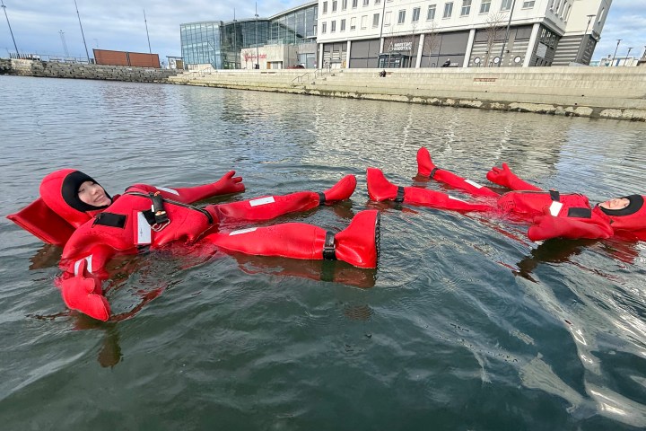Two people in red suits floating in water near a building.