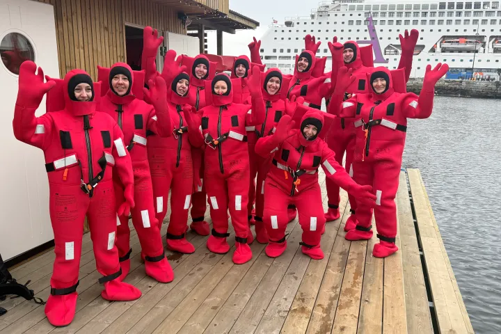 Group in red survival suits on dock, cruise ship in background.