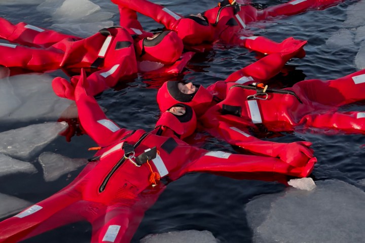 People in red survival suits floating on icy water, surrounded by ice chunks.
