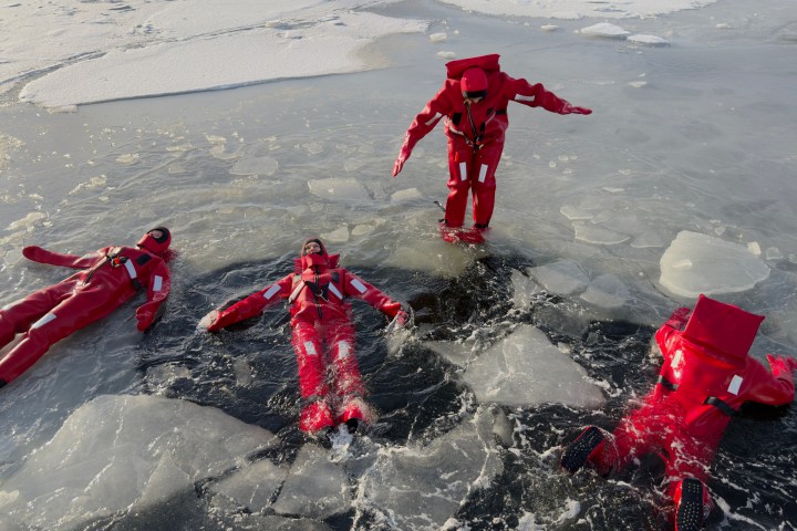 Four people in red suits lying and standing on icy water with floating ice chunks.