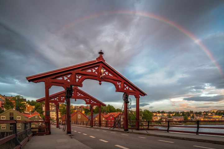Wooden bridge with arched red beams under a rainbow, cloudy sky.