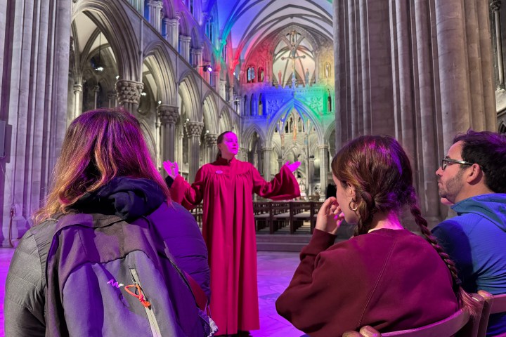 Woman in red gown speaks to three seated people in a cathedral with colorful lighting.